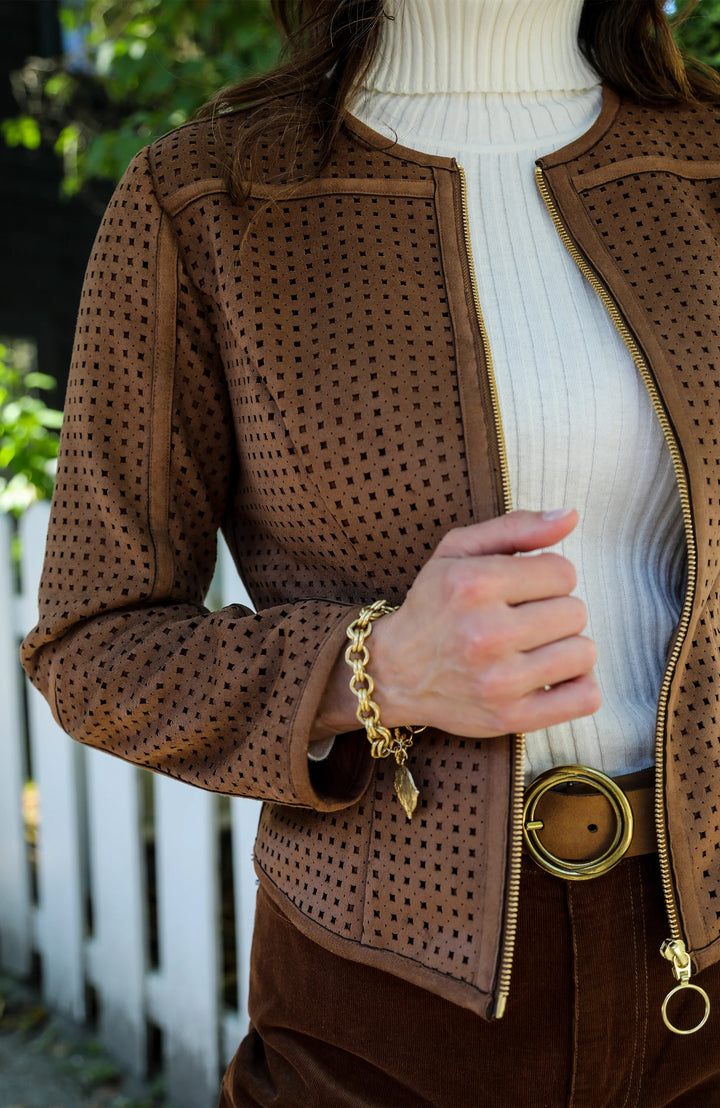 Close-up of gold ring-pull zipper and contrast piping on the punched faux suede jacket, worn over a white turtleneck with a gold chain bracelet and belt.