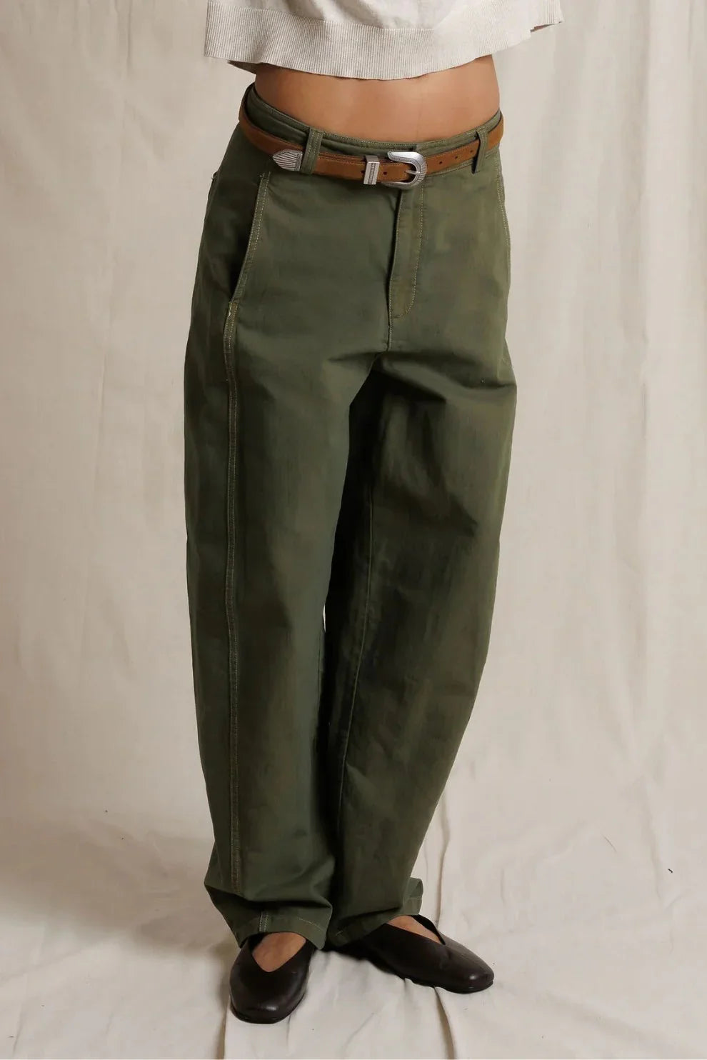 Close-up front view of a model wearing dark olive barrel-leg utility pants with a brown leather belt and silver buckle, paired with a cream top and black ballet flats.