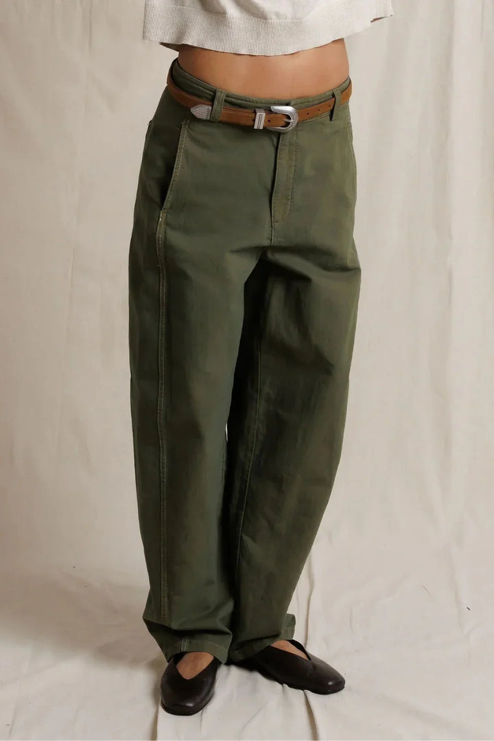 Close-up front view of a model wearing dark olive barrel-leg utility pants with a brown leather belt and silver buckle, paired with a cream top and black ballet flats.