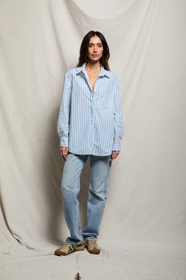 Woman wearing blue and white stripe oversized cotton poplin button-down shirt open over white tank, half-tucked into light-wash straight-leg denim, styled with tan sneakers, neutral studio backdrop — perfectwhitetee Avalon Shirt