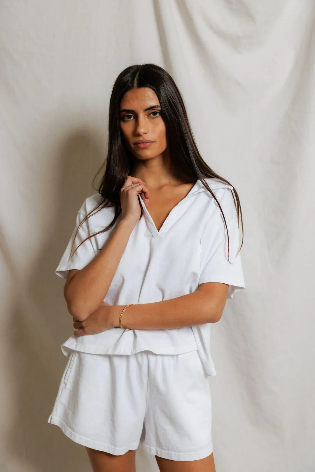 Woman wearing white cropped boxy polo with open V-notch collar and matching white shorts, styled with gold bangle bracelet, neutral studio backdrop — perfectwhitetee Charlie Polo