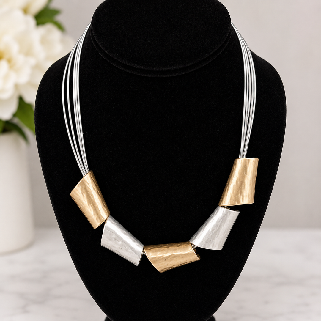 Necklace with gold and silver geometric pendants on a black stand with a blurred floral background