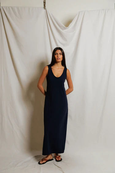 Woman wearing navy cotton maxi tank dress with scoop neckline and wide shoulder straps, hands in pockets, styled with black flip sandals, neutral studio backdrop — perfectwhitetee Jasmine Dress