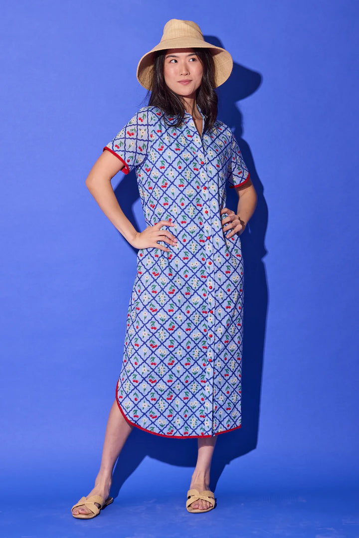Alden Adair Josie Midi Dress in light blue cherry-and-floral lattice print with red contrast trim, shown front on-model, button-front placket, short collar, short sleeves, relaxed midi silhouette with side slits, styled with woven sandals and a straw sun hat.