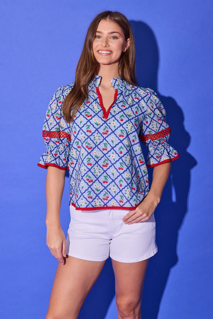 Alden Adair Lottie Top in light blue cherry-and-daisy lattice print with red crochet trim, shown front on-model, split V-neckline with ruffled stand collar, puff sleeves with crochet-inset band and ruffled cuffs, relaxed body, styled with white shorts.