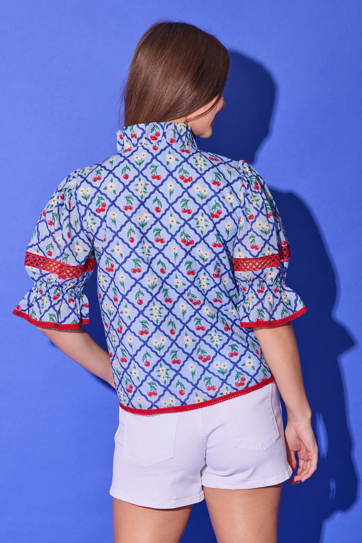 Alden Adair Lottie Top, back view in light blue cherry-and-daisy lattice print, showing the ruffled stand collar, puff sleeves with red crochet inset bands and ruffled cuffs, and red crochet trim at the hem.