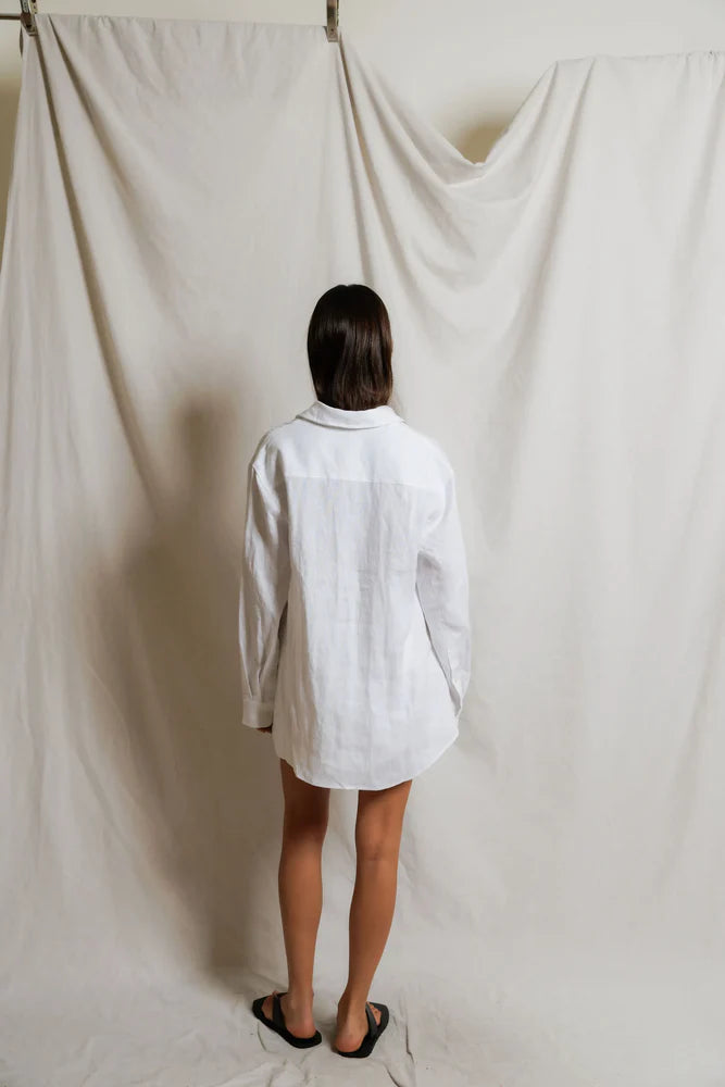 Model wearing Perfectwhitetee Lorelai Linen Oversized Button-Down in white, styled open over matching linen shorts with black flat sandals, against draped cream backdrop. - back