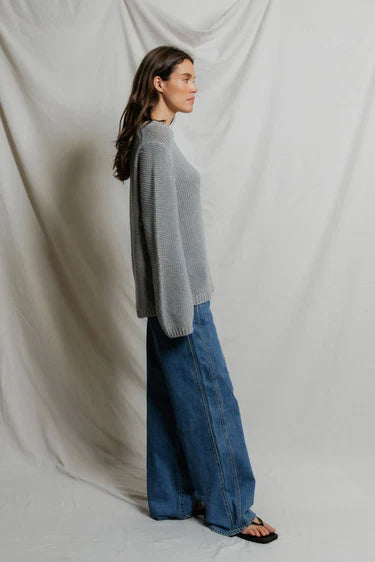 Model wearing Perfectwhitetee Margaret Cotton Slouchy Open-Knit Sweater in heather grey, styled with wide-leg blue denim, shown from the back at a three-quarter turn, against draped grey backdrop. - side