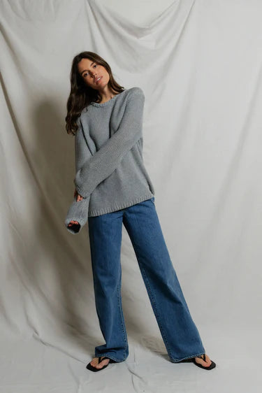 Model wearing Perfectwhitetee Margaret Cotton Slouchy Open-Knit Sweater in heather grey, styled with wide-leg blue denim, shown from the back at a three-quarter turn, against draped grey backdrop.