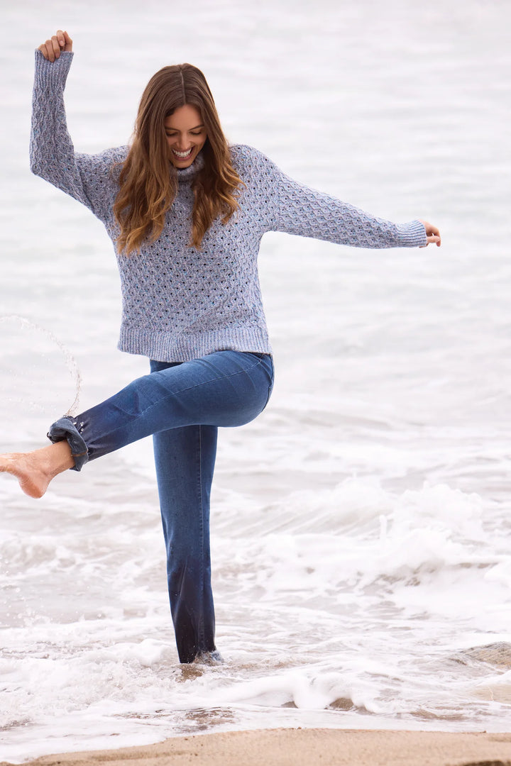 Full-body beach photo of a model kicking one foot into the surf, wearing a light blue cable-knit turtleneck sweater and cuffed straight-leg cropped jeans.