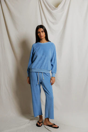 Woman wearing Pacific blue loop terry capri pant with matching terry pullover, natural drawcord waistband visible, styled with black flip sandals, neutral studio backdrop — perfectwhitetee Eliana Pant