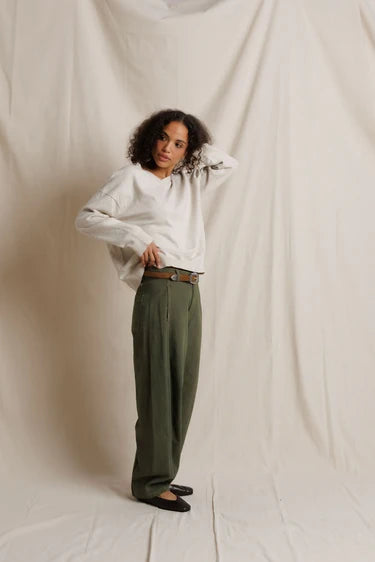 Full-body shot of a model with curly hair wearing the dark olive utility pants with a cream sweater and black ballet flats, one hand resting in her hair.