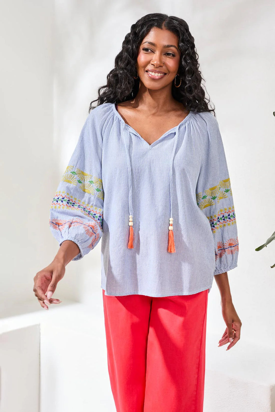 Model wearing Tribal Raglan Balloon Sleeve Blouse in baja blue stripe with embroidered balloon sleeves and beaded tassel neckline tie, styled with wide-leg red trousers and tan heeled sandals.